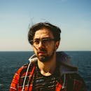 A man wearing glasses and a plaid jacket stands by the sea in Istanbul.