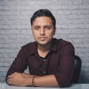 Confident adult Indian man in trendy shirt sitting at white table near brick wall while looking at camera in light room